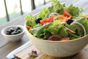 Saladeira com salada de alface verde crocante, tomates vermelhos, cebola roxa e cenoura ralada sobre mesa de madeira clara com ingredientes frescos ao fundo