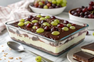 Bombom de uva na travessa de vidro com camadas de creme branco, uvas inteiras verdes e roxas e cobertura de chocolate brilhante