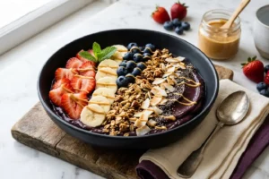 Tigela de açaí fit cremosa em bowl de cerâmica preta com granola dourada, frutas vermelhas frescas e fio de pasta de amendoim sobre mármore claro